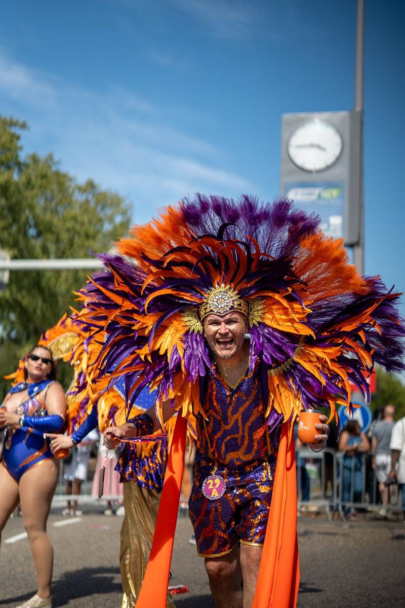 Een lachende man met een enorme tooi van oranje en paarse veren loopt op de camera af.